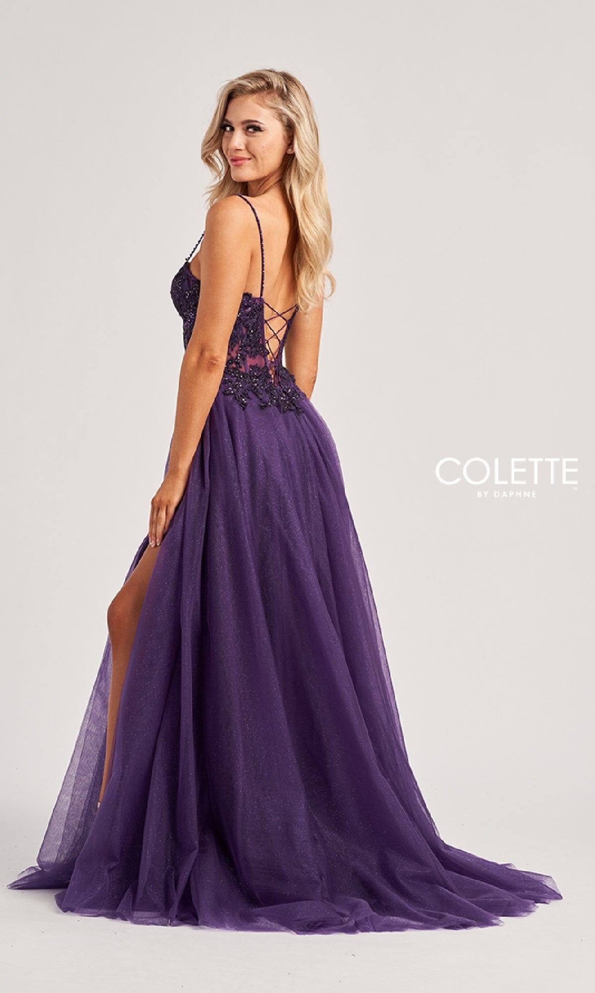 A woman in the Colette CL8280 long prom dress—a floor-length purple gown with a lace-up back and side slit—stands looking over her shoulder. The
